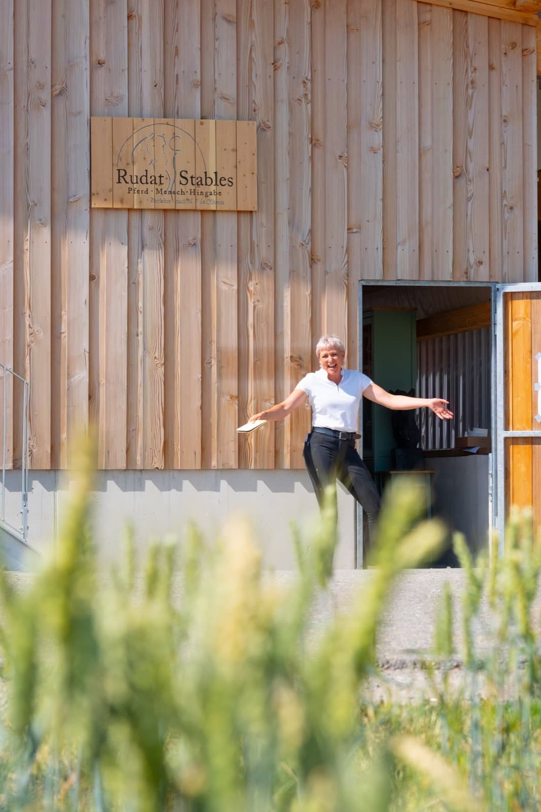 Vor dem Schild „Rudat Stables“ mit dem Zusatz „Pferd · Mensch · Hingabe“ am Stallgebäude breitet eine Frau lachend die Arme aus