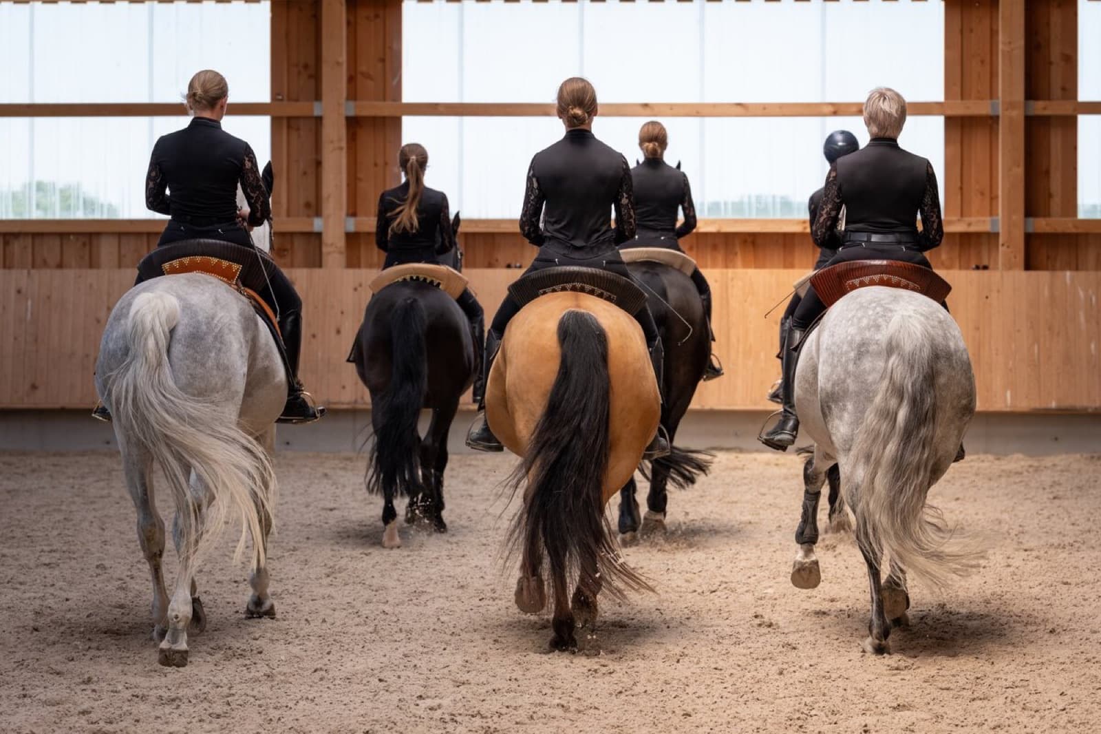 Fünf Reiterinnen in schwarzer Kleidung reiten nebeneinander von der Kamera weg durch die Halle
